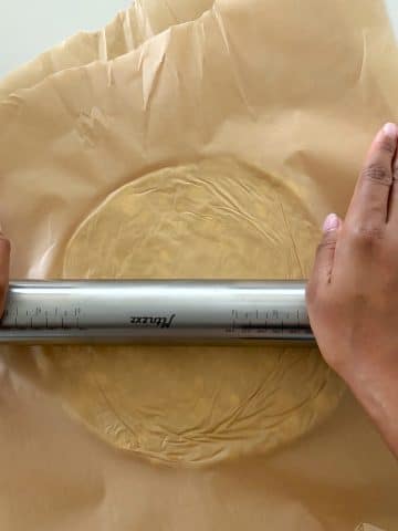 Two hands using a rolling pin to roll out pie dough in between sheets of parchment paper.