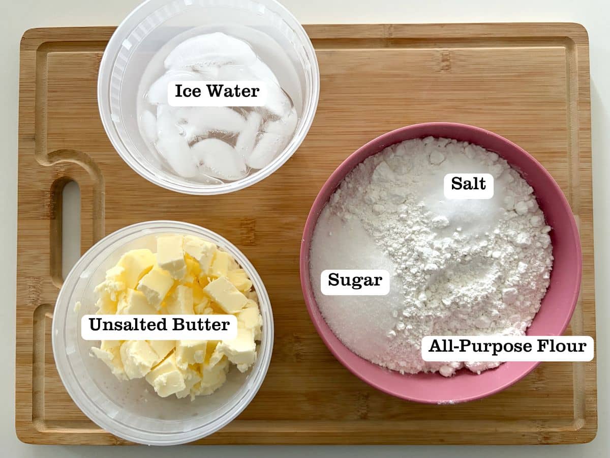 A board with containers of ingredients to make pie dough - including butter, ice water, and flour.