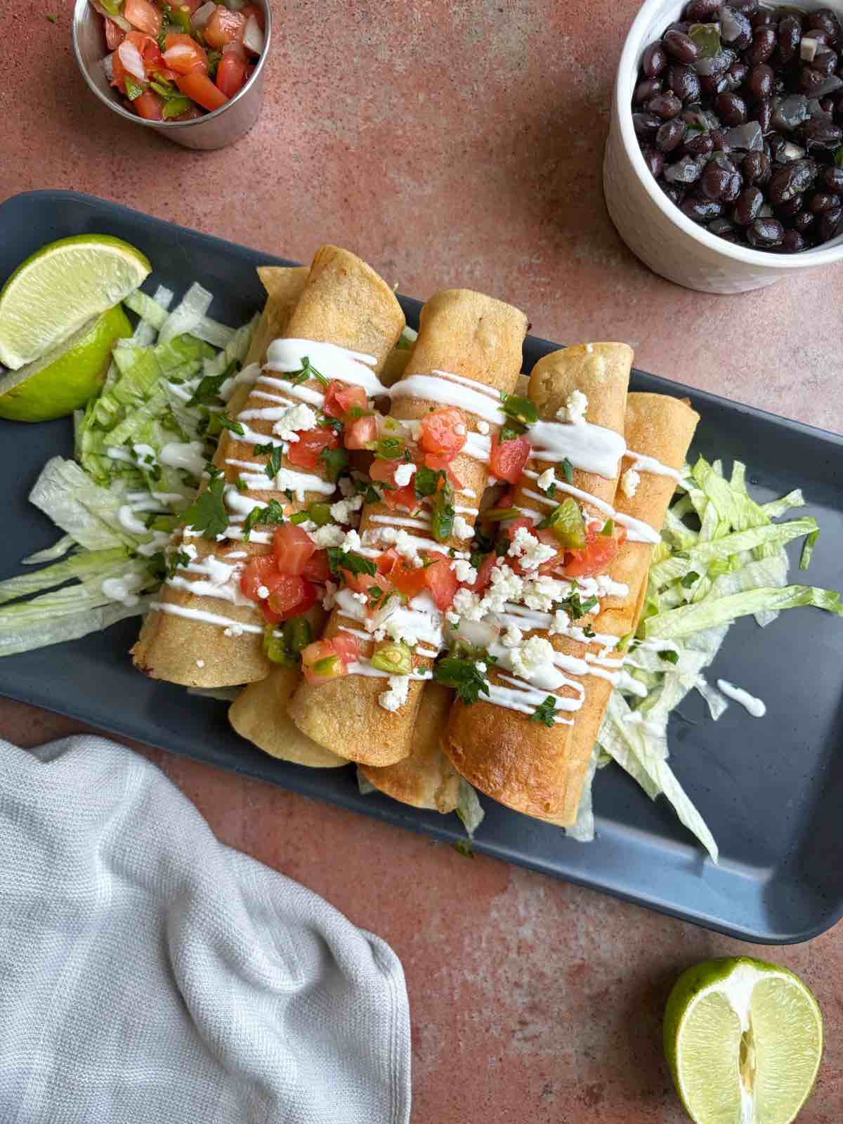 A tray of chicken tinga taquitos with accompaniments like lettuce and pico de Gallo on top.