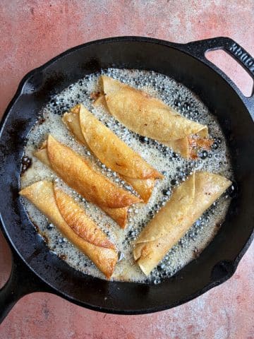 Rolled taquitos frying in a cast iron skillet filled with oil.