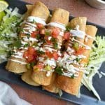 A stack of crispy chicken taquitos over shredded lettuce, with sour cream and pico de Gallo on top.
