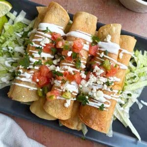 A stack of crispy chicken taquitos over shredded lettuce, with sour cream and pico de Gallo on top.