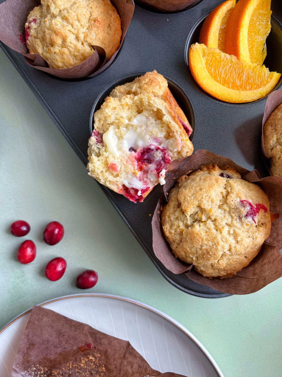A muffin tin with cranberry orange muffins, one halved and slathered in butter.