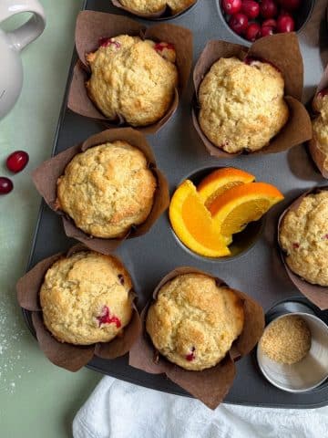 A muffin pan filled with baked cranberry orange muffins, and some ingredients in a few of the cups.