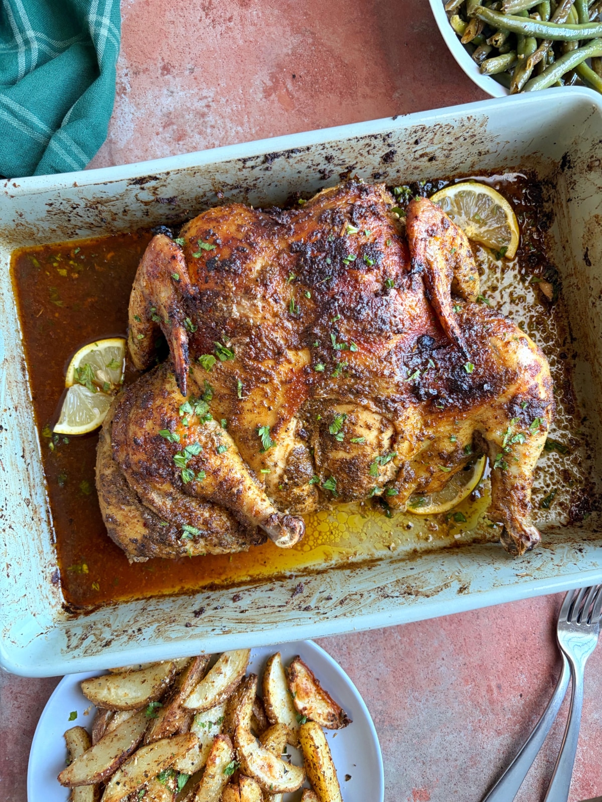 A whole roasted spatchcock chicken with roasted potatoes and green beans in bowls on the side.